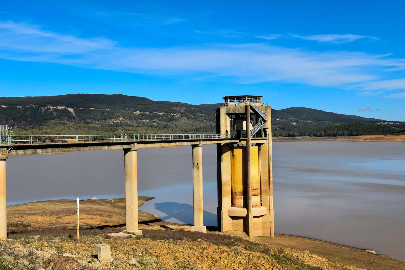 A concrete water intake tower linked by a bridge walkway to the shore of a reservoir, hills in the background.